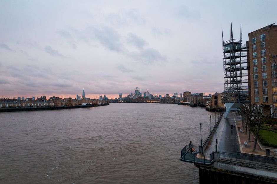 A wide view of a river in an urban setting during twilight, with calm, slightly rippled water extending from the foreground toward the distant skyline. On the right side, there is a modern metal obser